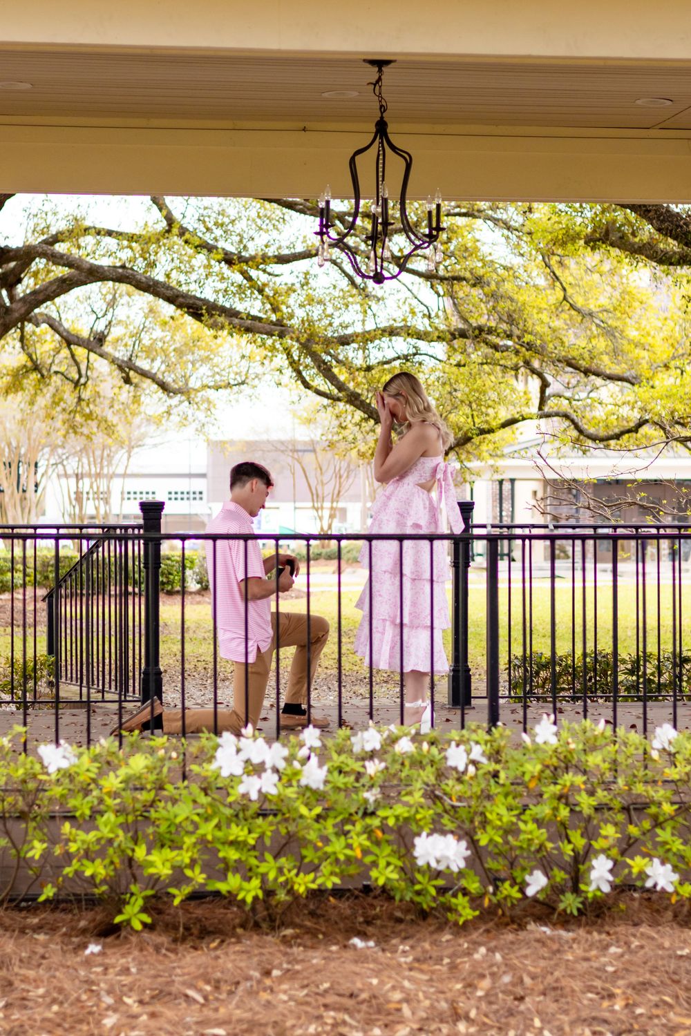 Man proposing on one knee to woman in pink dress on garden porch with white flowers and iron fence.