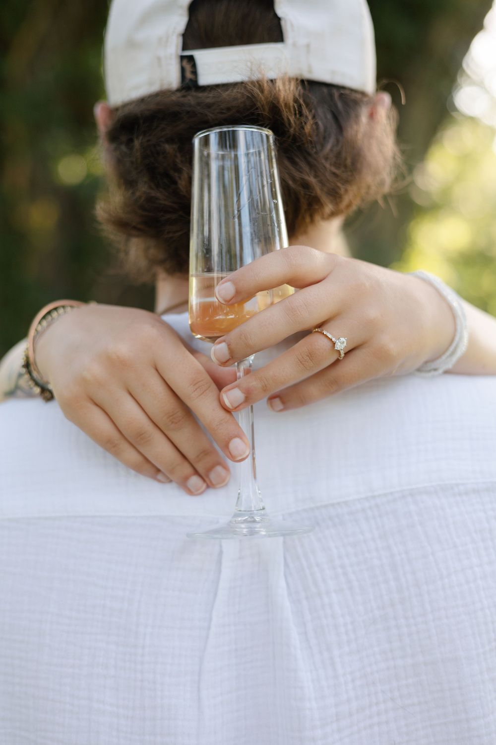 Woman holding champagne flute while embracing partner, showcasing diamond engagement ring and bracelet outdoors.