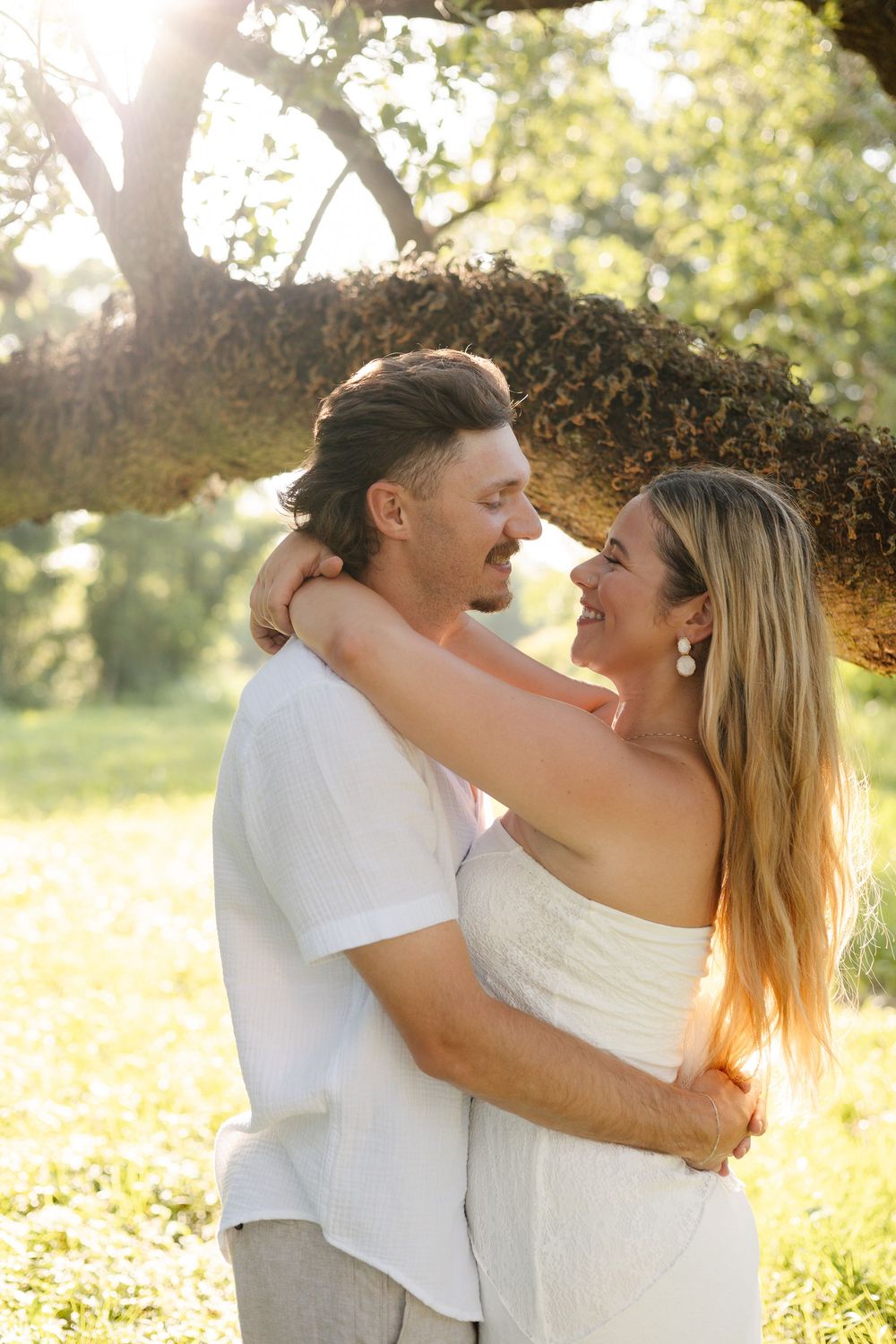 A couple embraces and smiles at each other under a sunlit tree in a lush green park setting.
