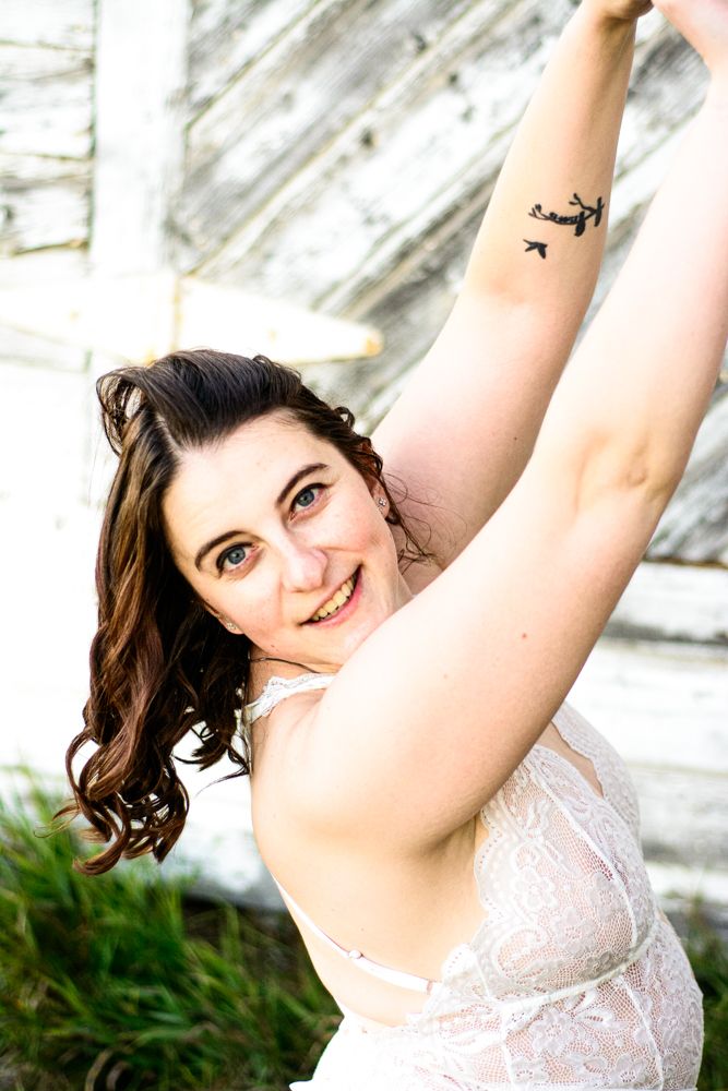 Portrait in white lace top with small airplane tattoo visible on arm against weathered wood background.