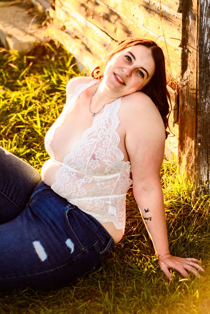 Casual outdoor portrait in lace top and jeans sitting by weathered barn wall in golden sunlight.