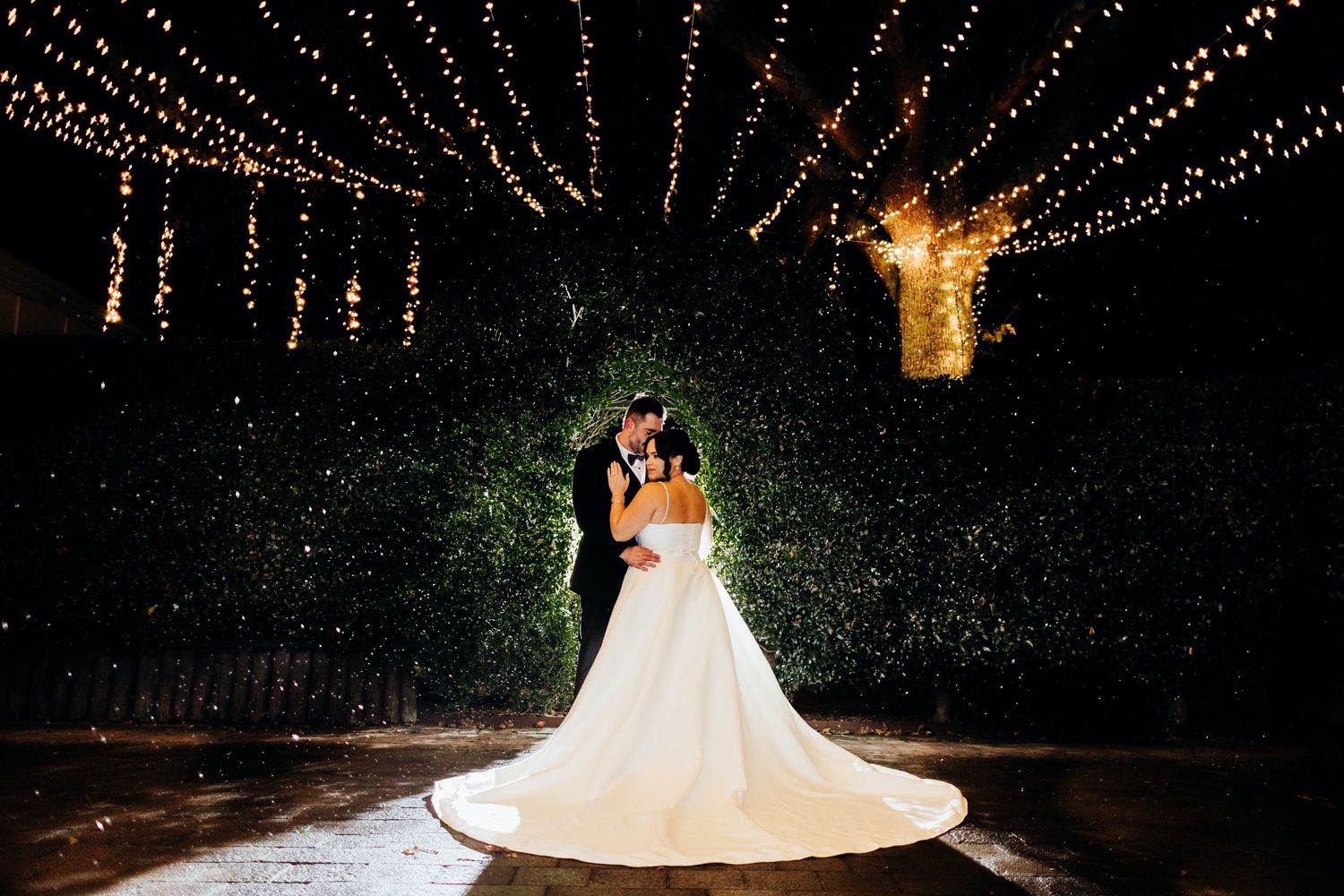 Romantic evening wedding portrait with twinkling string lights and lush greenery backdrop.