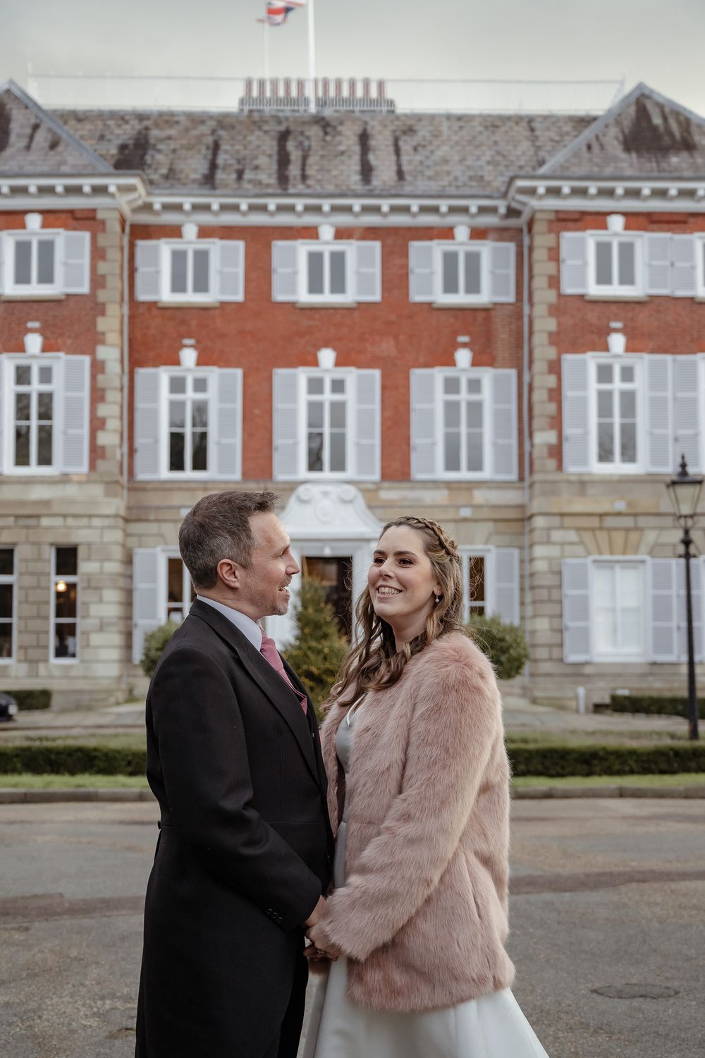 A couple stands close together in front of an elegant brick building with white trim.