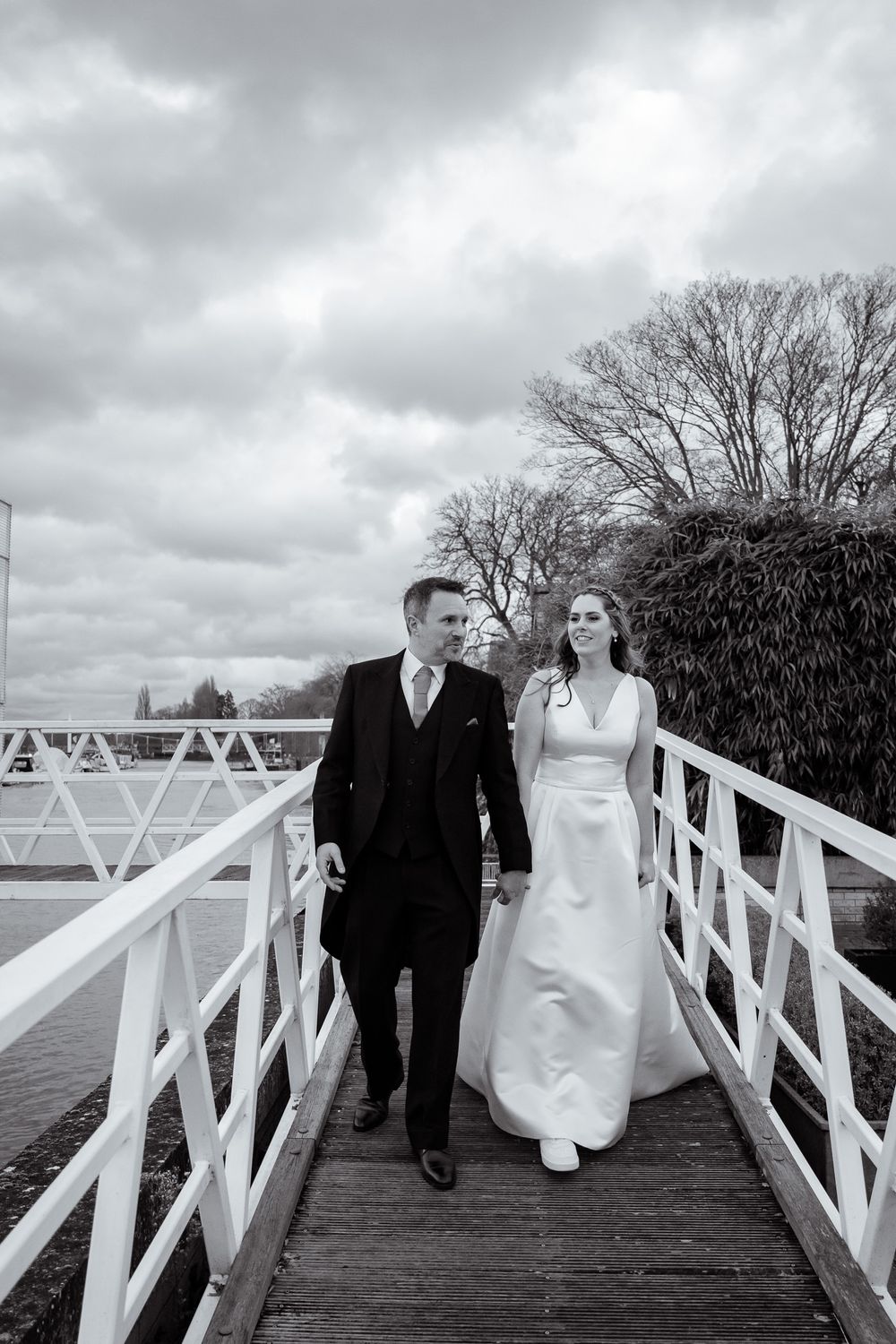 A couple walks along a white wooden bridge on their wedding day under dramatic cloudy skies.