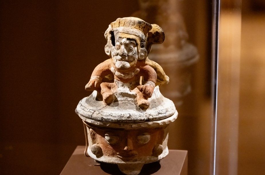 Clay vessel topped with an ornate decorative face or mask in ancient style.