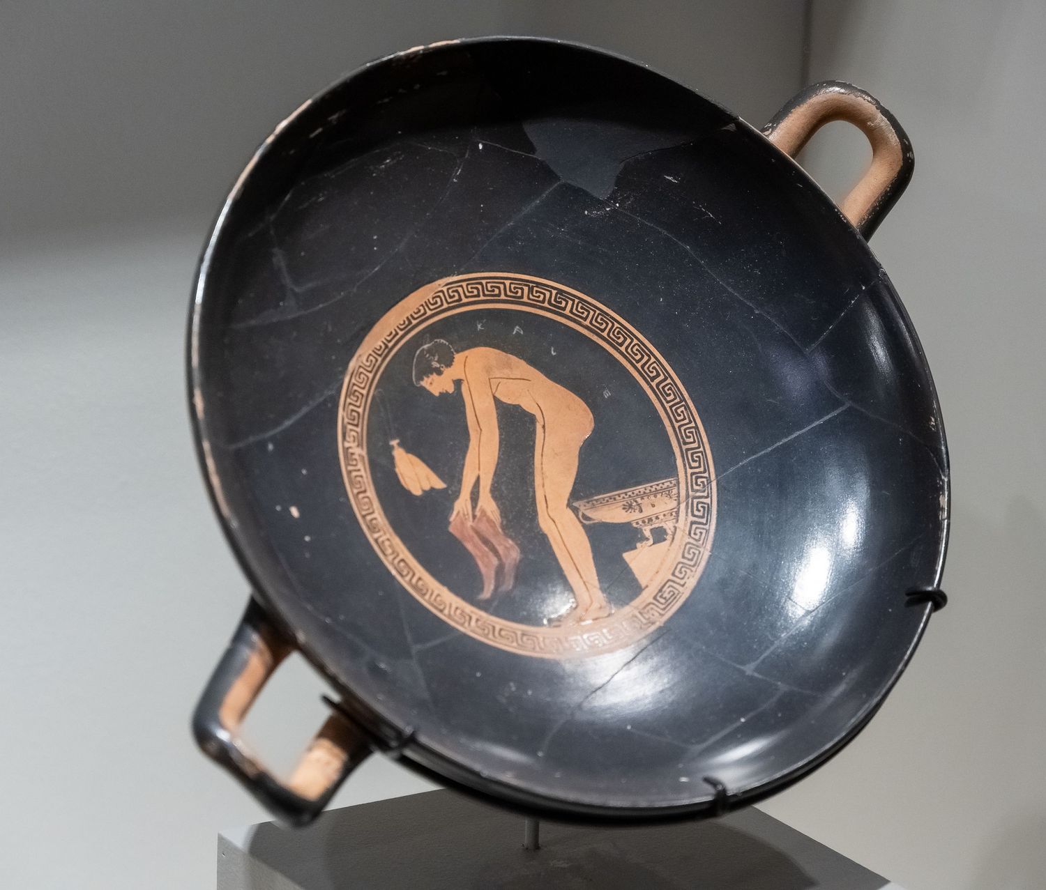 Black ancient Greek kylix bowl with red figure decoration and handles on display.