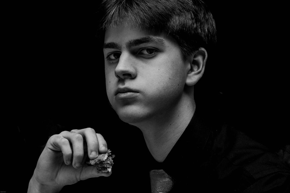 Black and white photo of a teenager holding a piece of a burger looking at the camera.