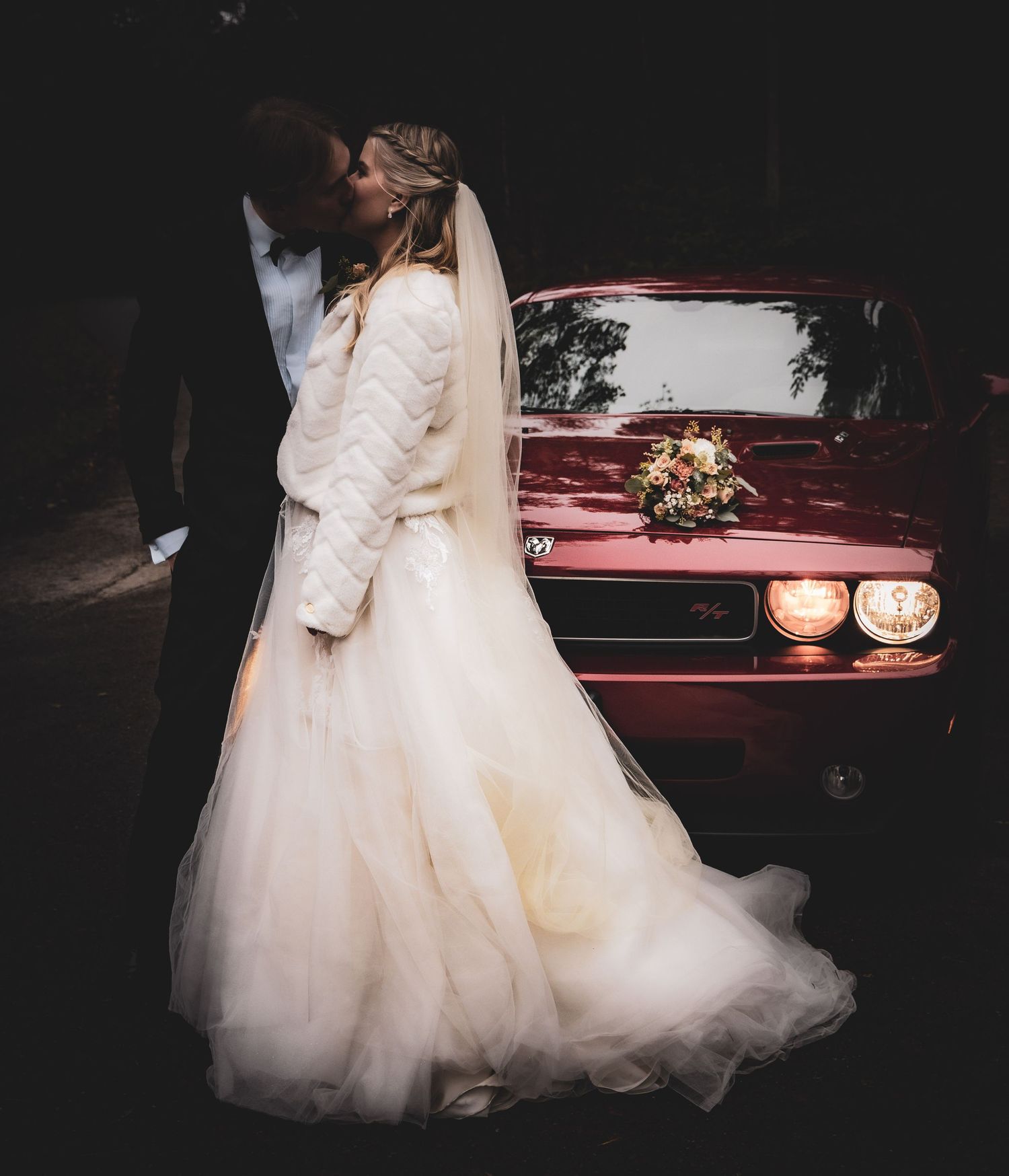 Bride and groom sharing a kiss beside a classic red Dodge Challenger at night.