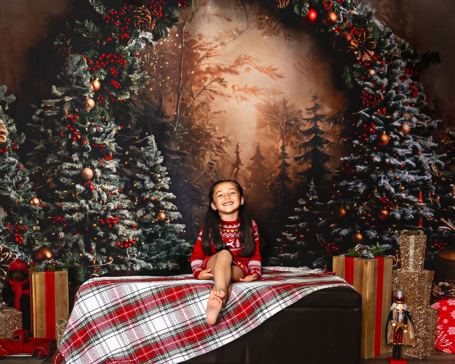 A festive Christmas photography setup with decorated evergreen trees, wrapped presents, and plaid blanket in warm lighting.