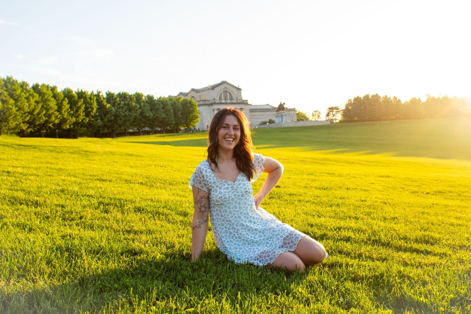 Someone in a polka dot dress sits in a sunlit meadow with a white house and trees in the background.