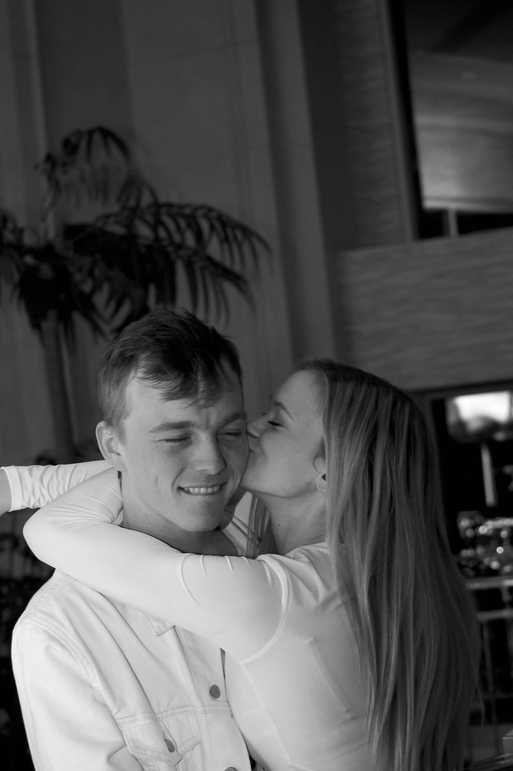 A couple shares an intimate moment as she kisses his cheek while he smiles in a black and white photo.