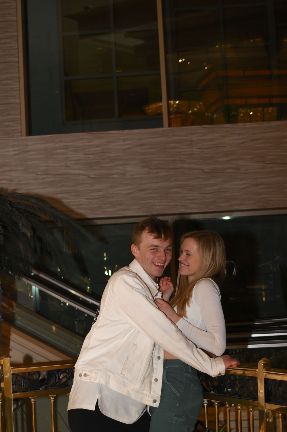 Two people in white clothing embracing and smiling on a balcony with city lights in the background.