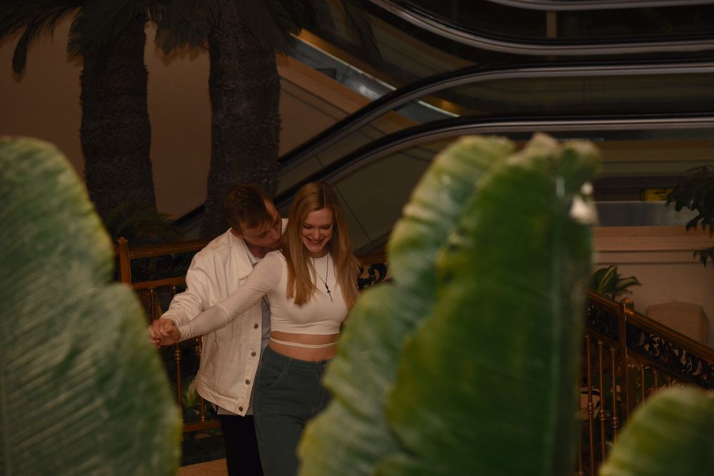 A couple in white clothing poses among large tropical plants in an indoor setting.