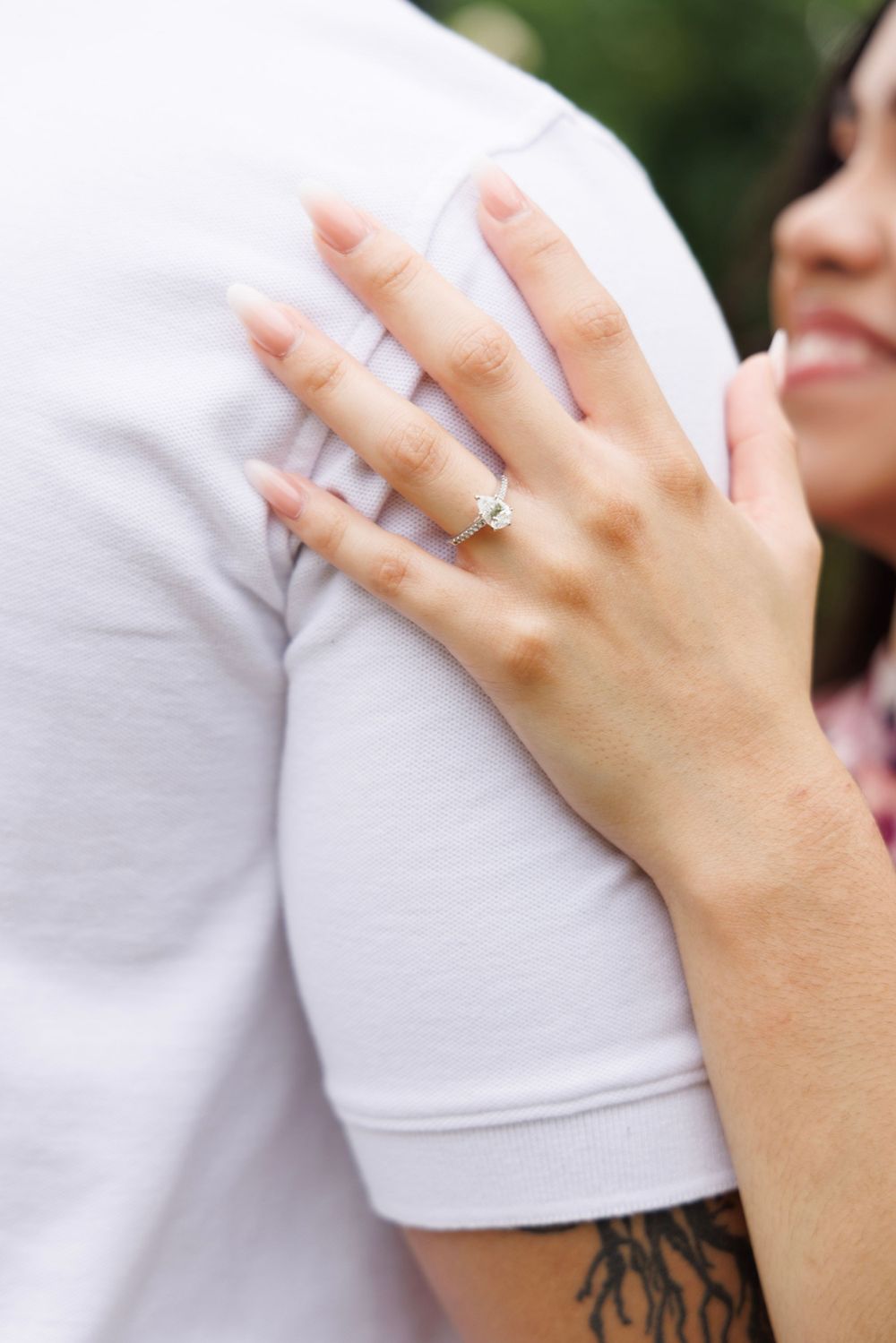 Close-up of a hand wearing an engagement ring resting on a white shirt with a visible tattoo.