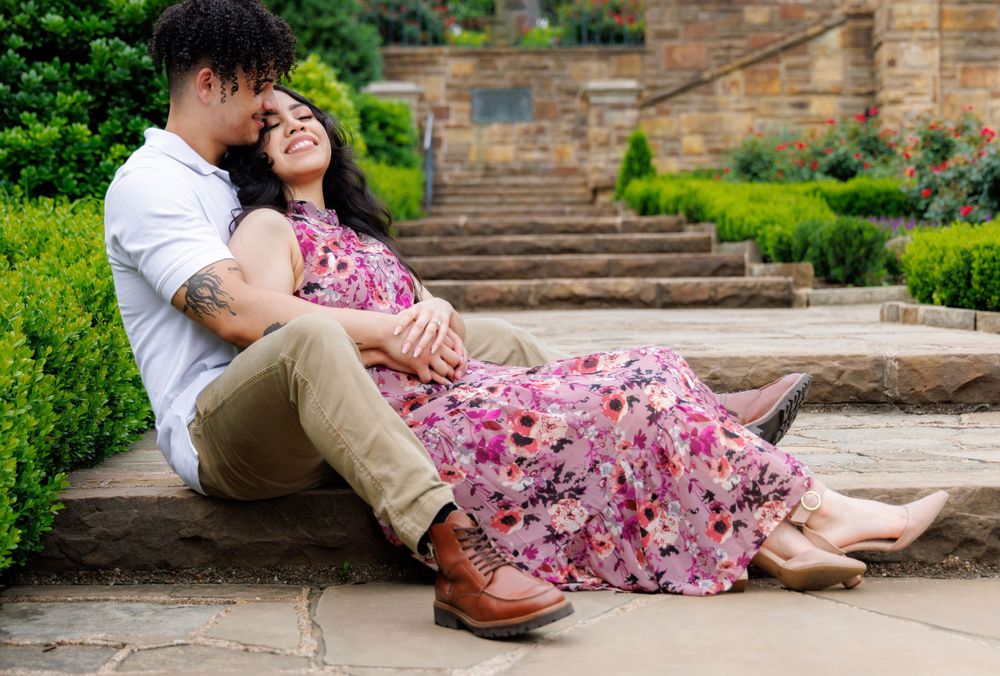 Couple relaxing on stone steps wearing white shirt and floral dress in garden setting.