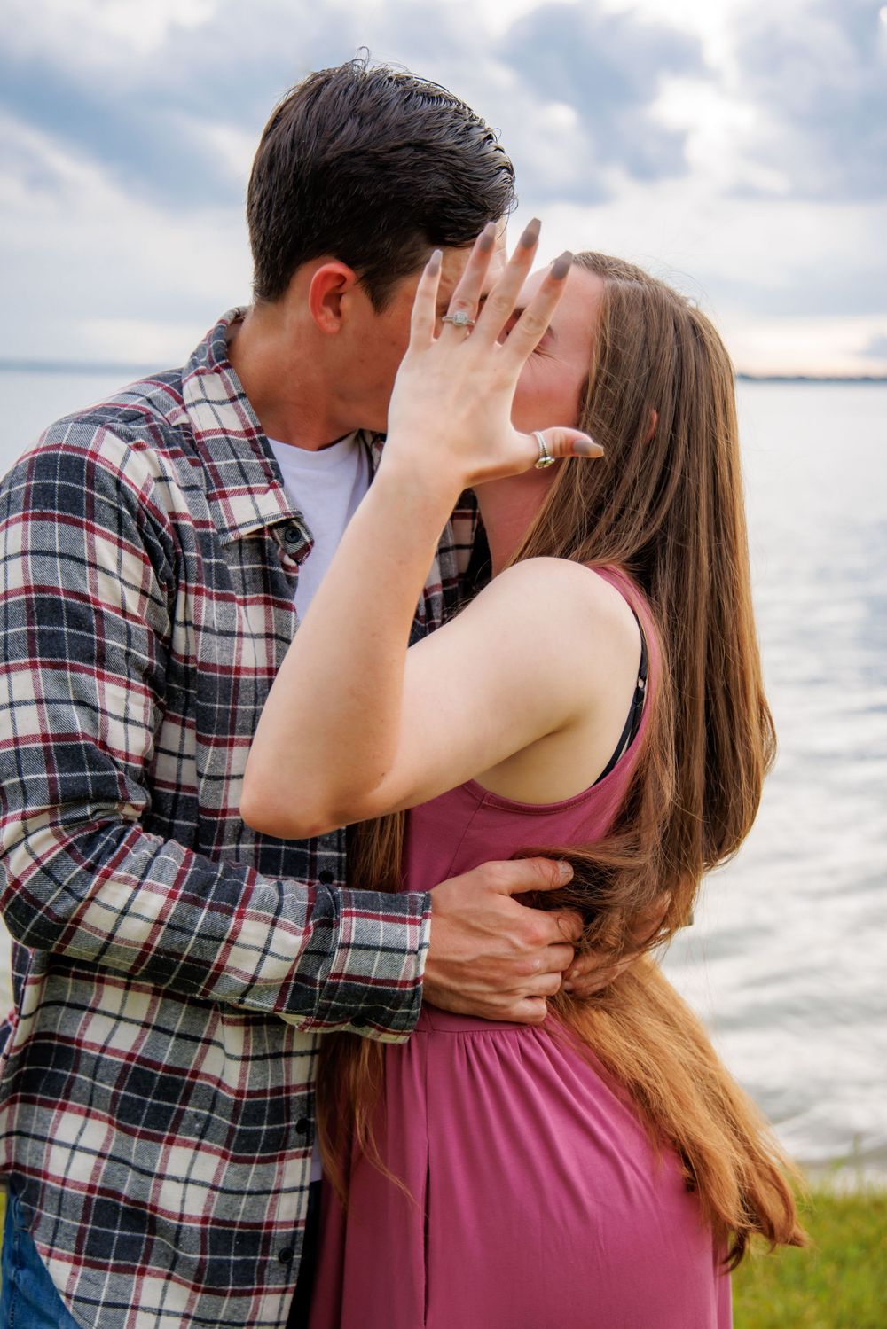 Couple sharing romantic moment by lake with woman showing engagement ring.
