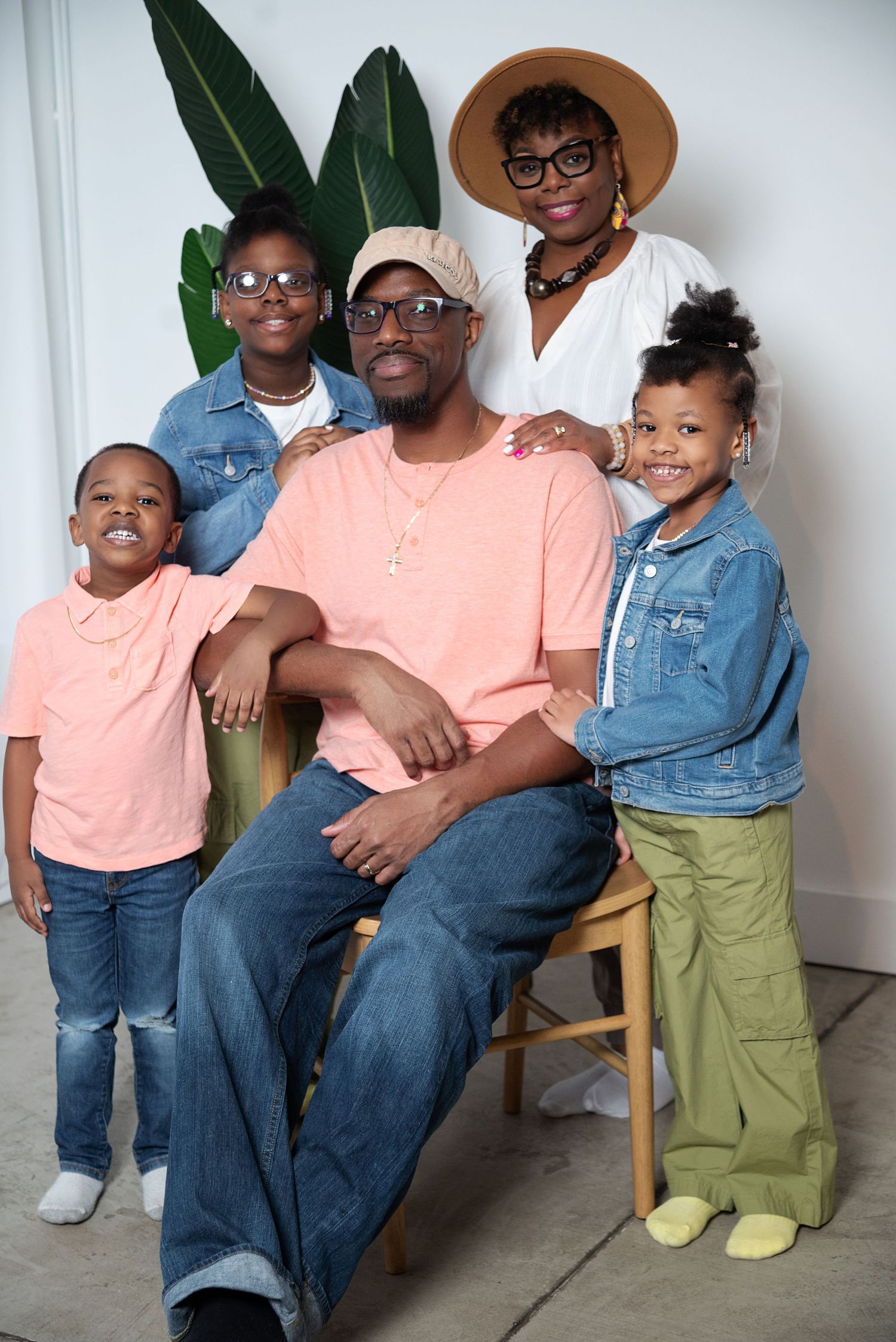 A family portrait showing a group of five in coordinated casual outfits with denim and pink tones against a white wall with plants.