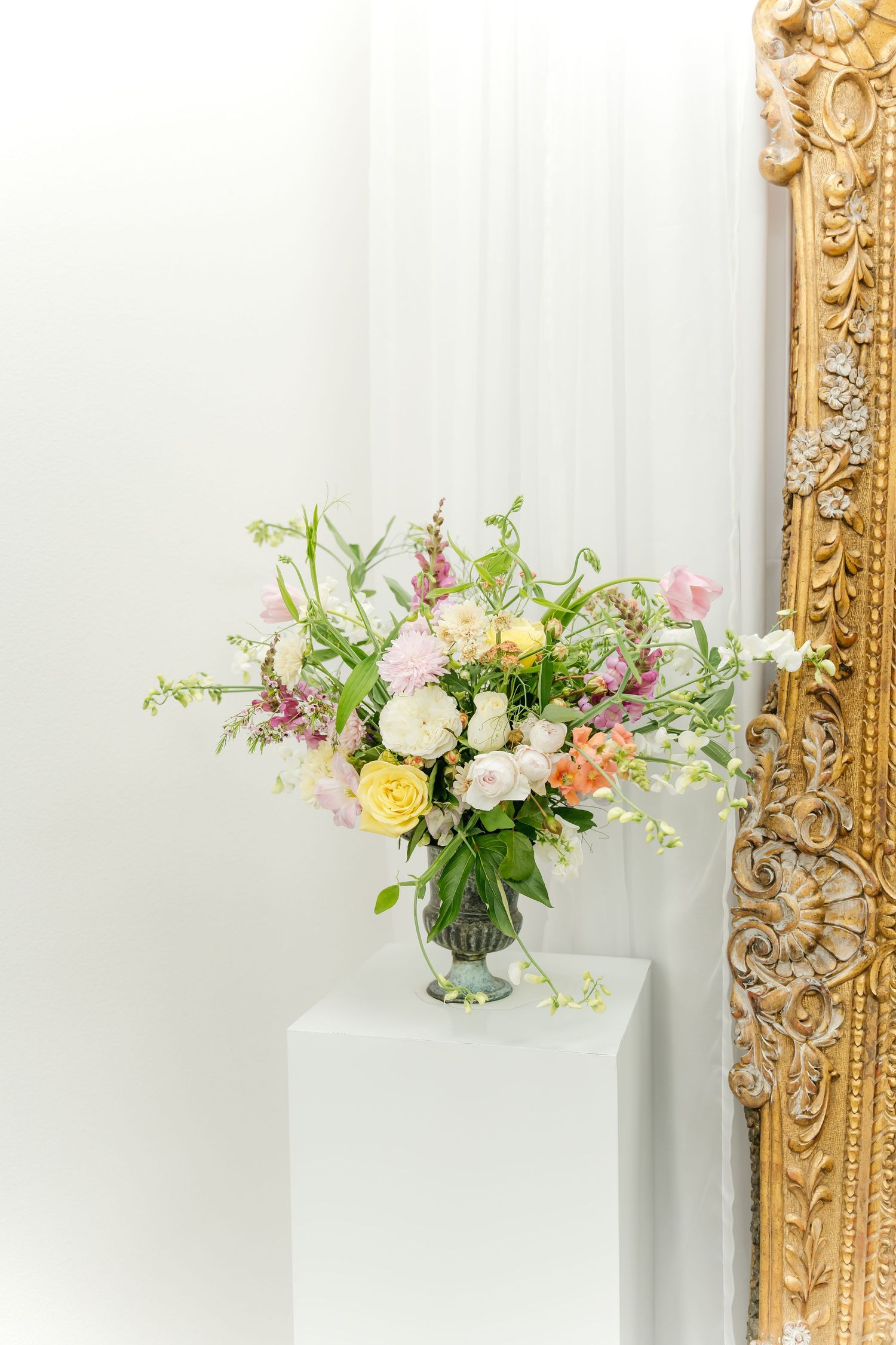 Elegant floral arrangements in glass vases sit atop tables with ornate gold-framed mirrors in a bright white room.