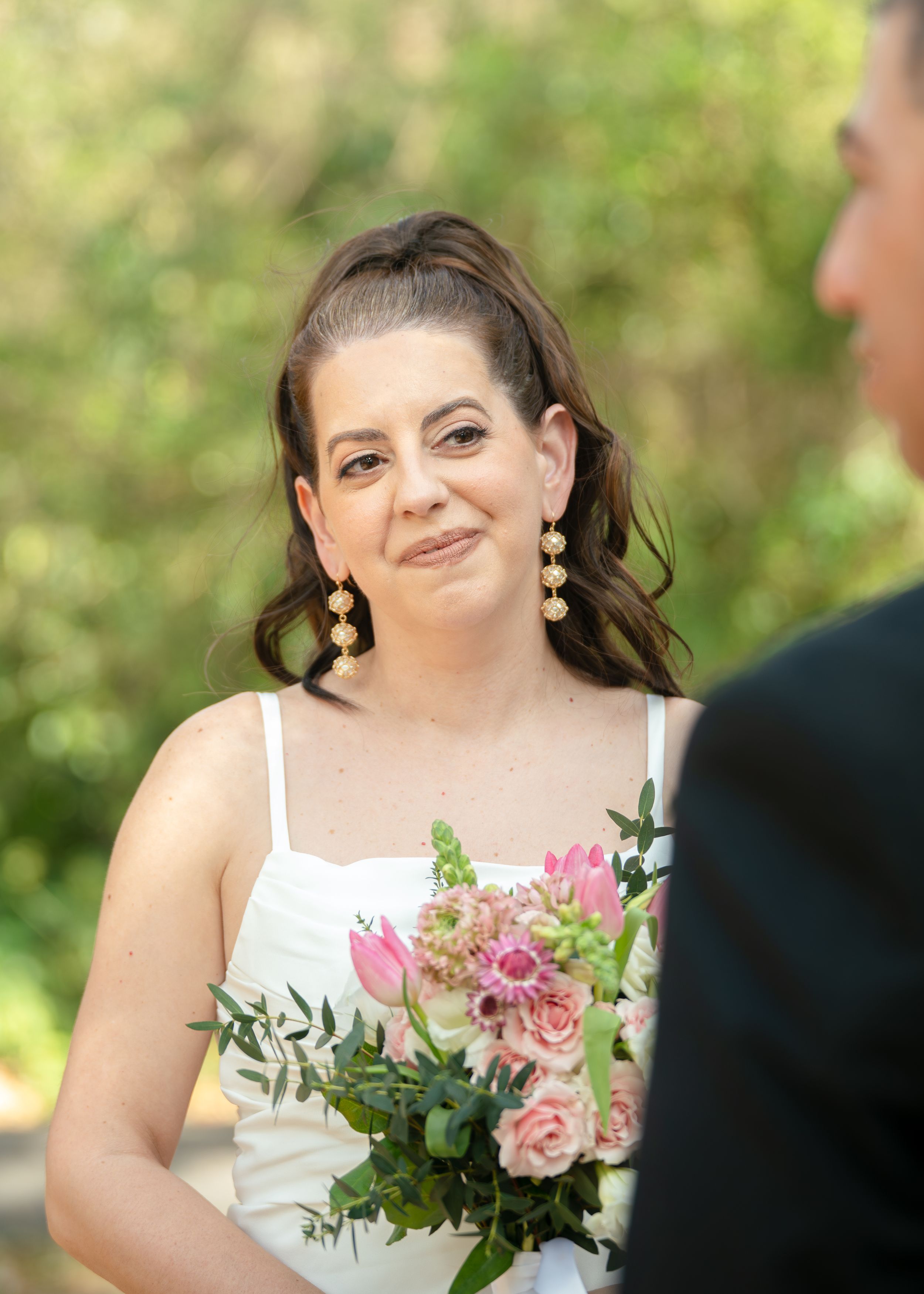 A bride in a strapless white lace wedding dress smiles while holding a bouquet of white flowers.