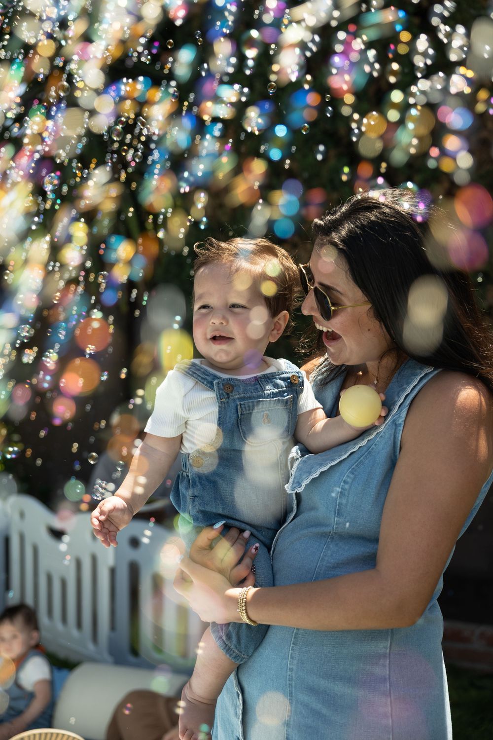 A mother in a denim dress holds her baby girl outdoors, surrounded by colorful floating soap bubbles in sunlight.