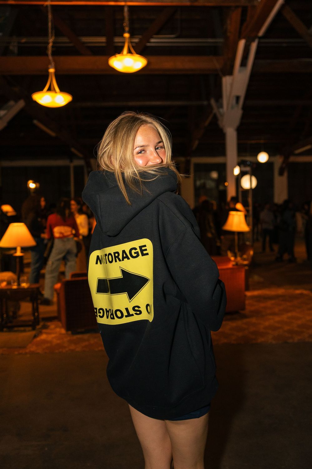 A person wearing a black hoodie with yellow text and arrow design stands in a dimly lit indoor space.