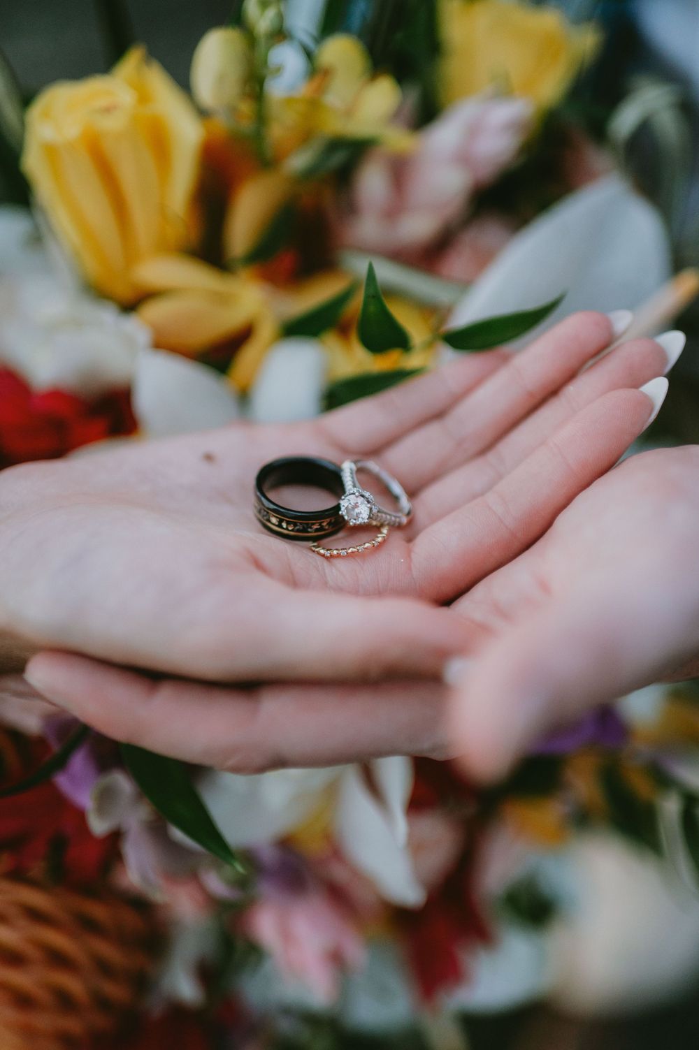 Wedding rings displayed on an open palm with colorful flowers in the background.