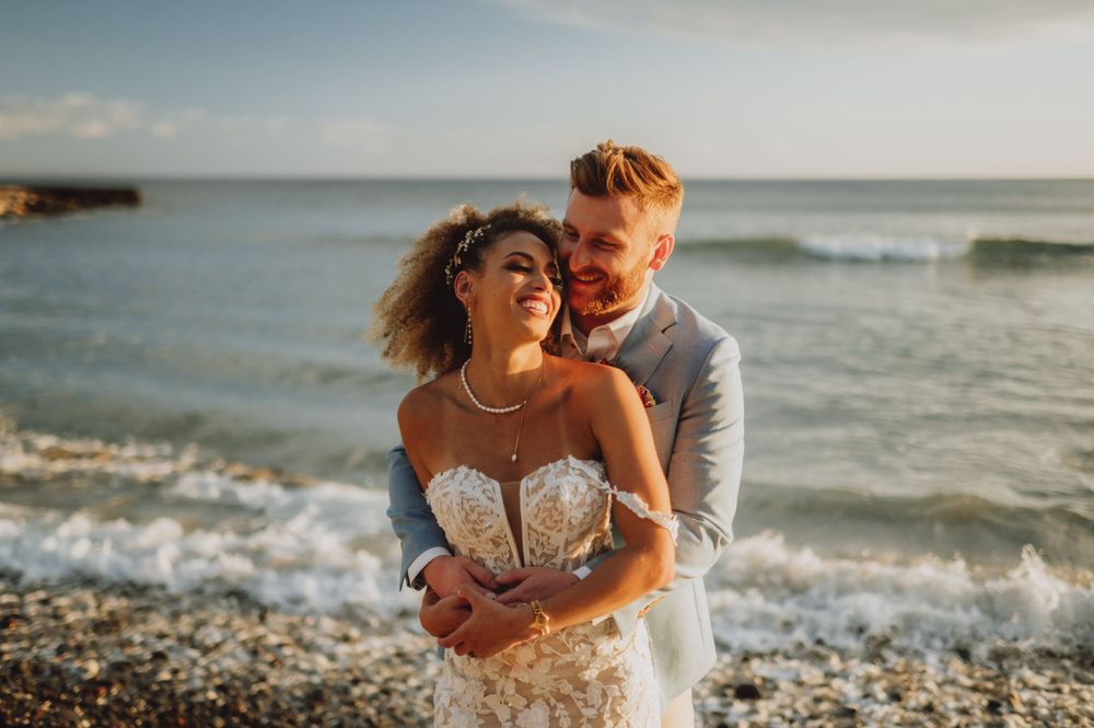 A romantic beach wedding photoshoot with waves crashing against the rocky shore at sunset.