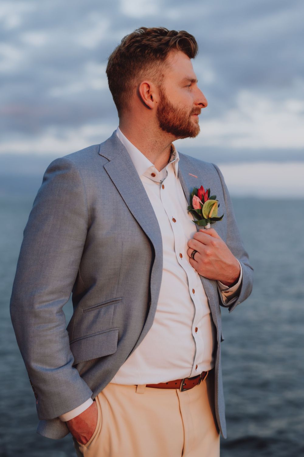 Groom in grey suit and khaki pants adjusts boutonniere against ocean backdrop.