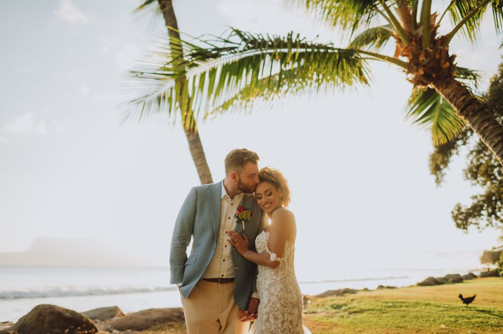 Wedding couple embracing under palm trees at sunset on tropical beach.