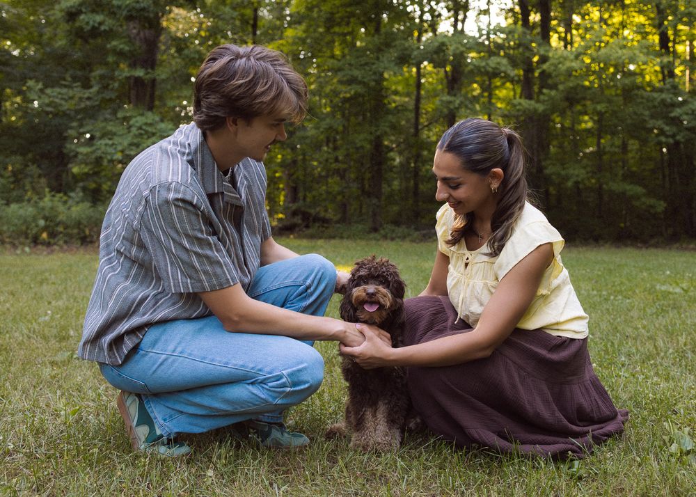 Two people crouching on grass petting a small brown curly-haired dog outdoors near trees.