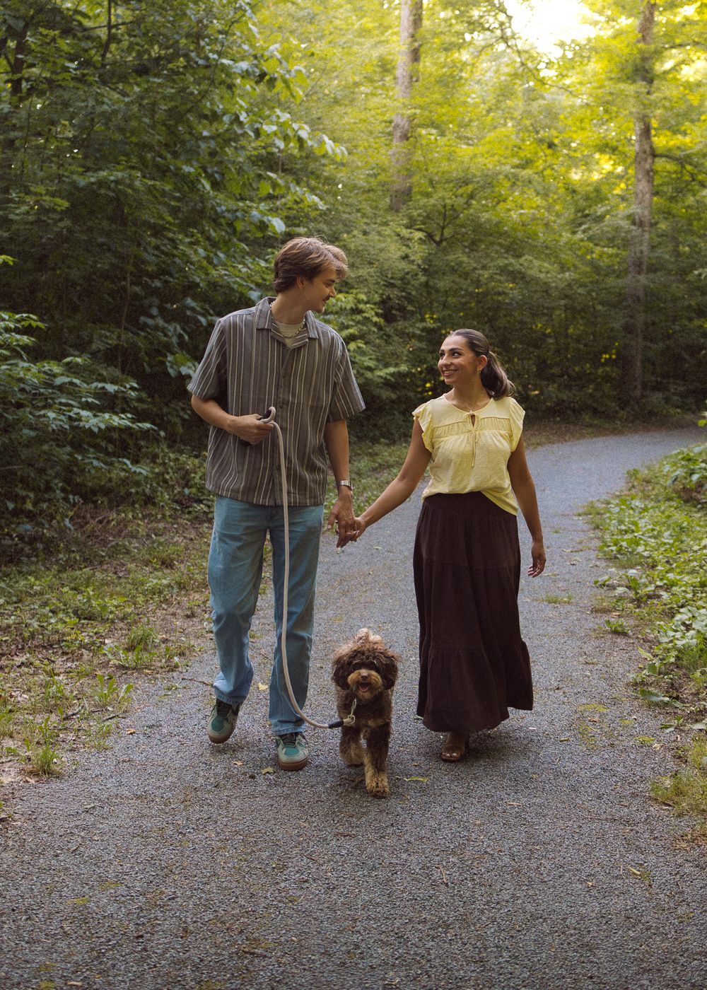 A couple walks hand-in-hand with a brown curly dog on a leash along a lush green forest path.