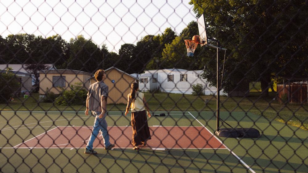 Two people playing basketball on an outdoor court, surrounded by a chain-link fence and green trees at sunset.