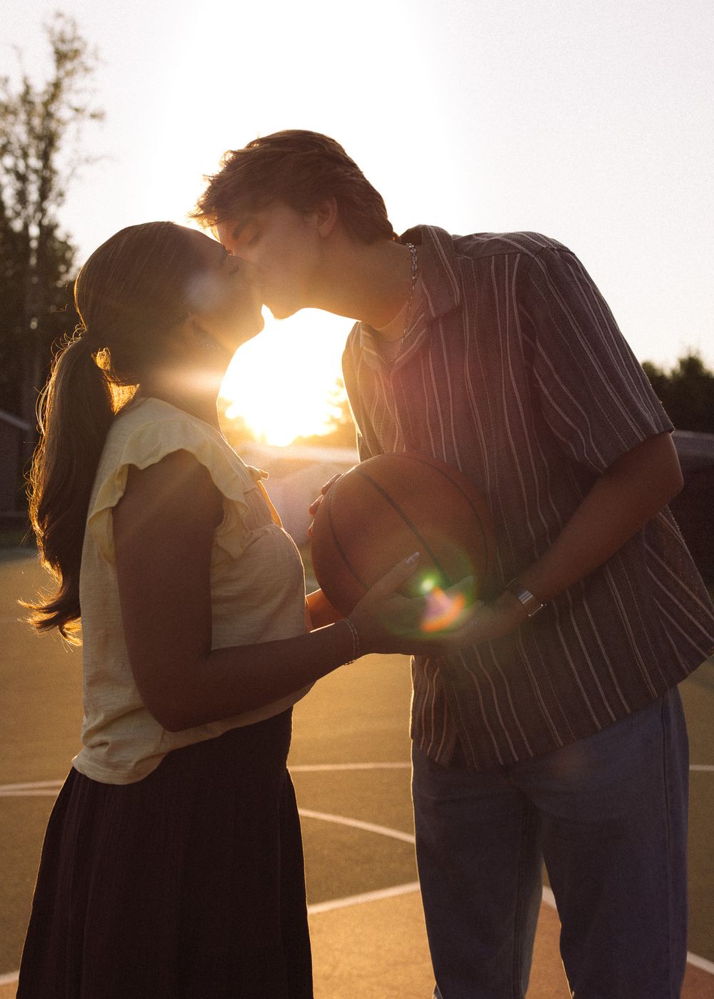 Couple sharing a romantic kiss at sunset on a basketball court, backlit by golden sun flare.