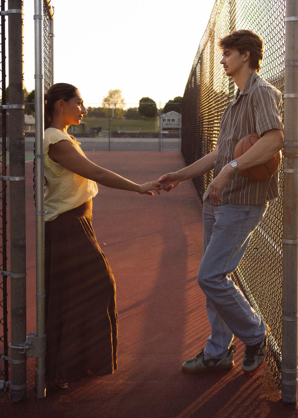 A young couple holds hands facing each other by a chain-link fence at golden hour on a tennis court.