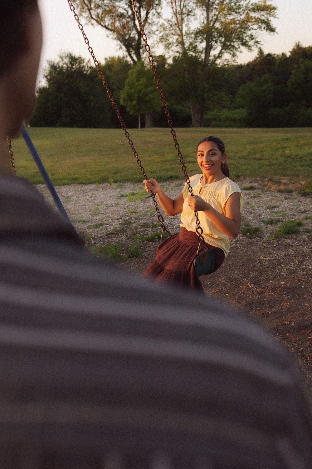 Young woman smiling on a swing set at golden hour in a park, holding chains while facing someone.