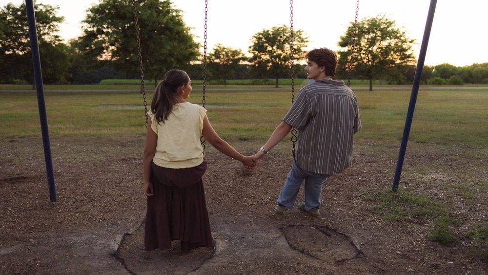 Couple holding hands at sunset near playground swings in a park, viewed from behind.