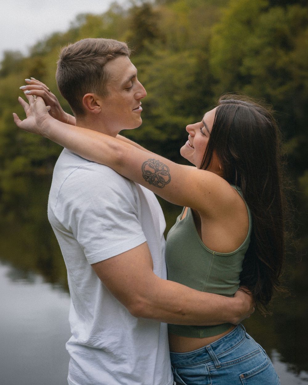 Couple embracing and smiling by a lake surrounded by lush green trees.