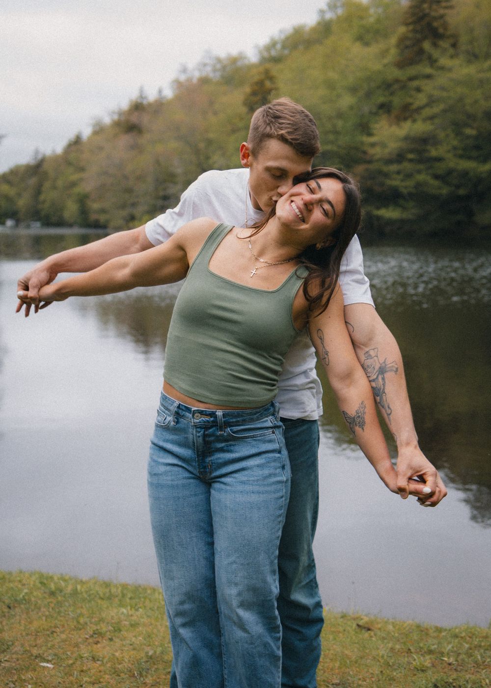 Man kissing womans cheek as she laughs with arms outstretched by a lake.