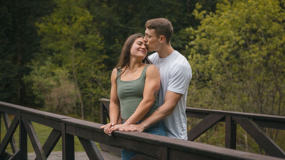 Romantic couple sharing a tender kiss on a wooden bridge surrounded by lush green forest trees.