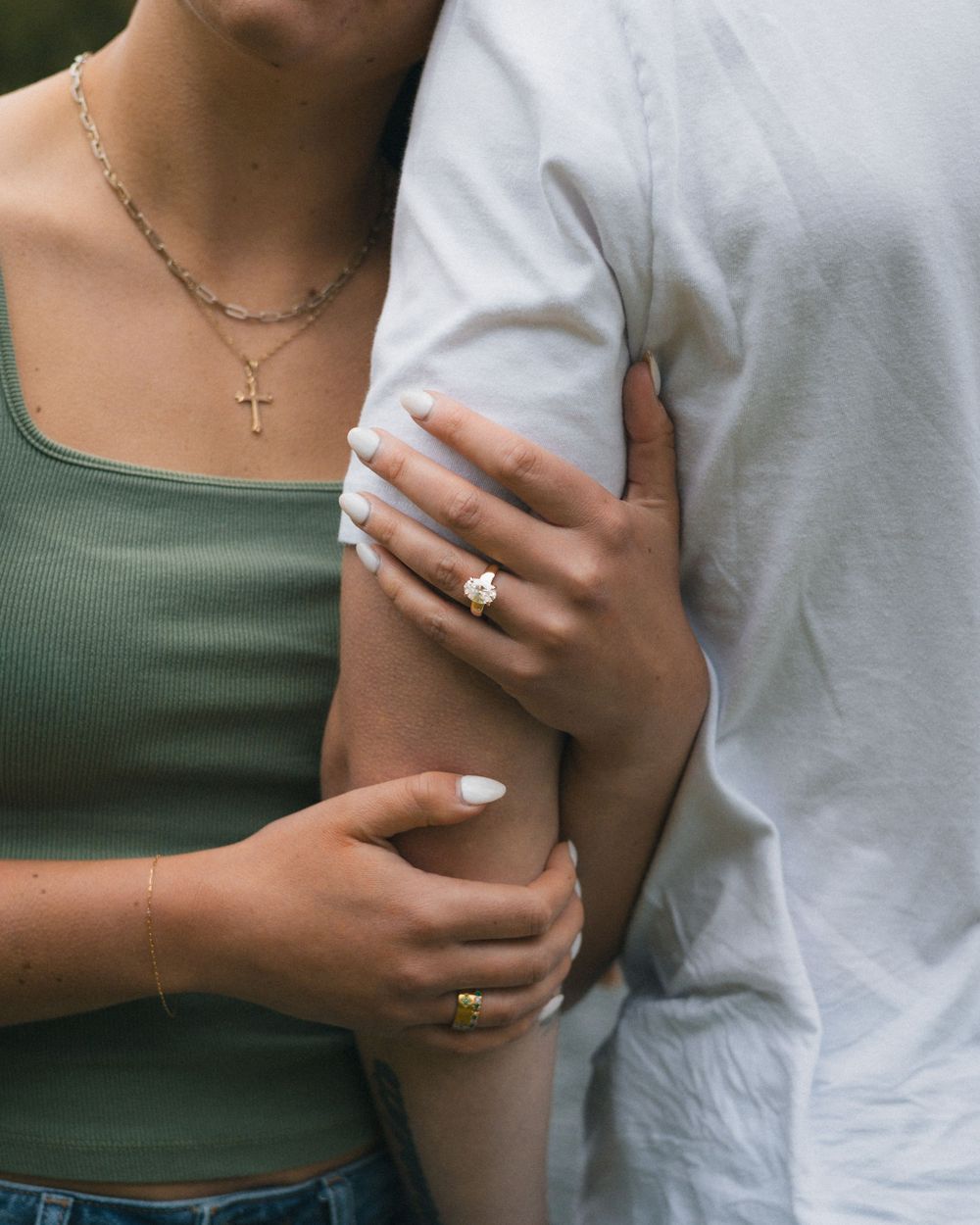 Couple embracing, woman wearing gold engagement ring, cross necklace, and white manicure, in sage green top.