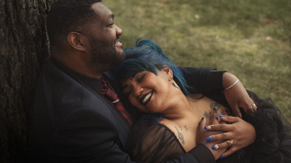 Couple laughing and embracing against a tree, woman with blue hair leaning on man in black suit outdoors.