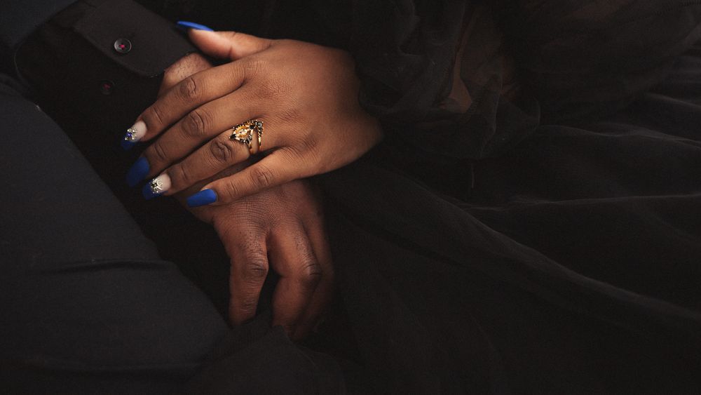 Close-up of hands with blue nails and a gold ring resting on dark fabric.