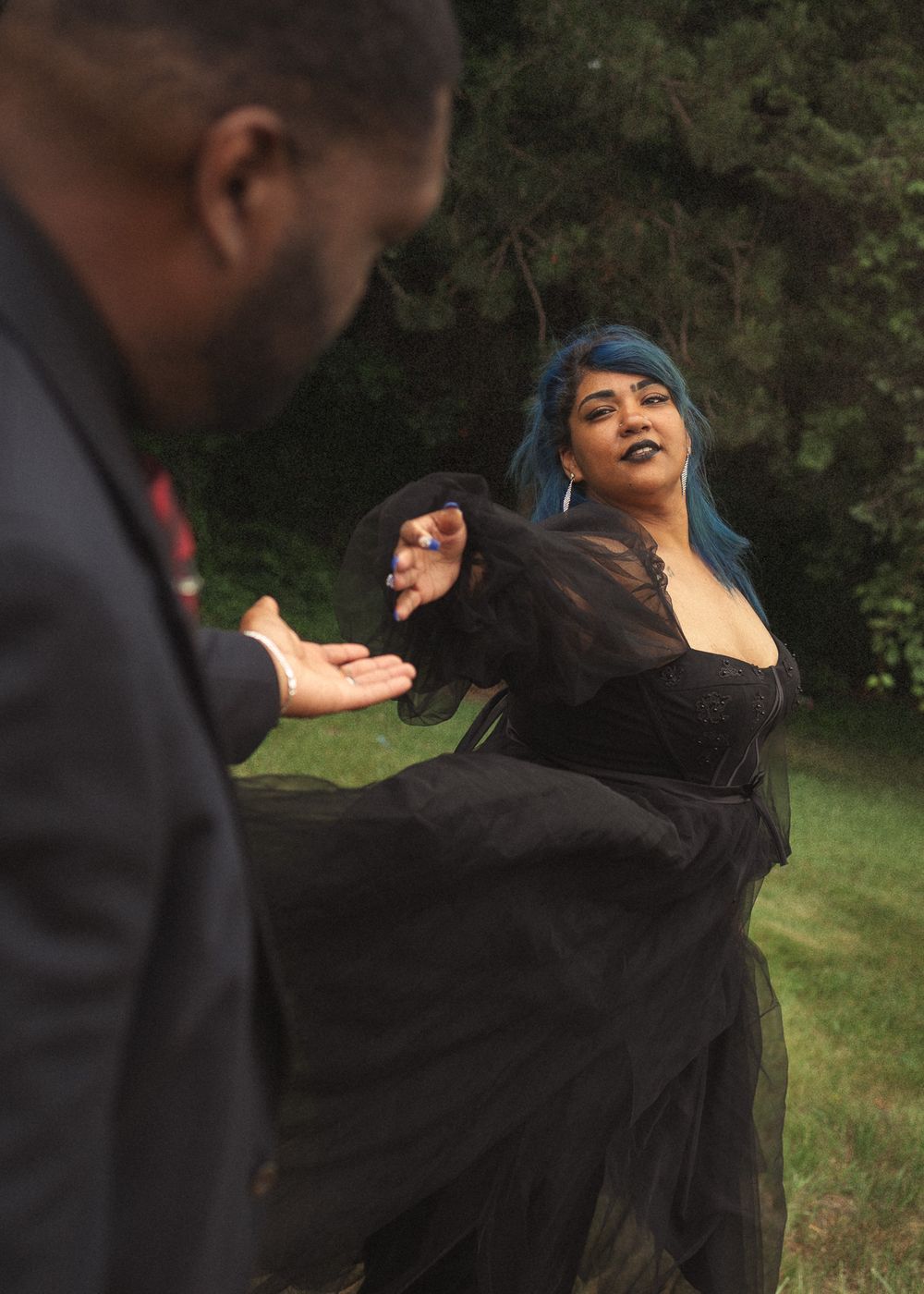 Woman in black gown with blue hair reaches toward man in suit outdoors.