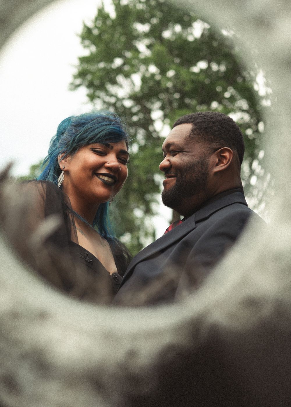 A smiling couple gazes at each other, woman with blue hair in black dress, man in dark suit, framed by a circular foreground element.