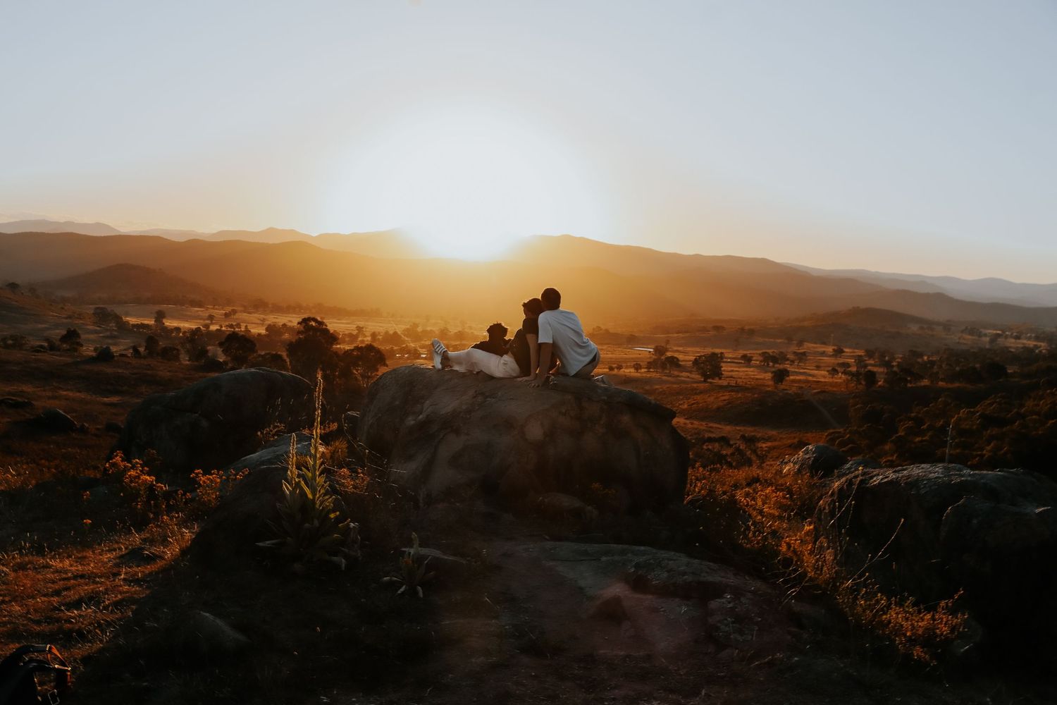 Couple sitting on rocks watching a golden sunset over a scenic valley landscape with mountains in the background.