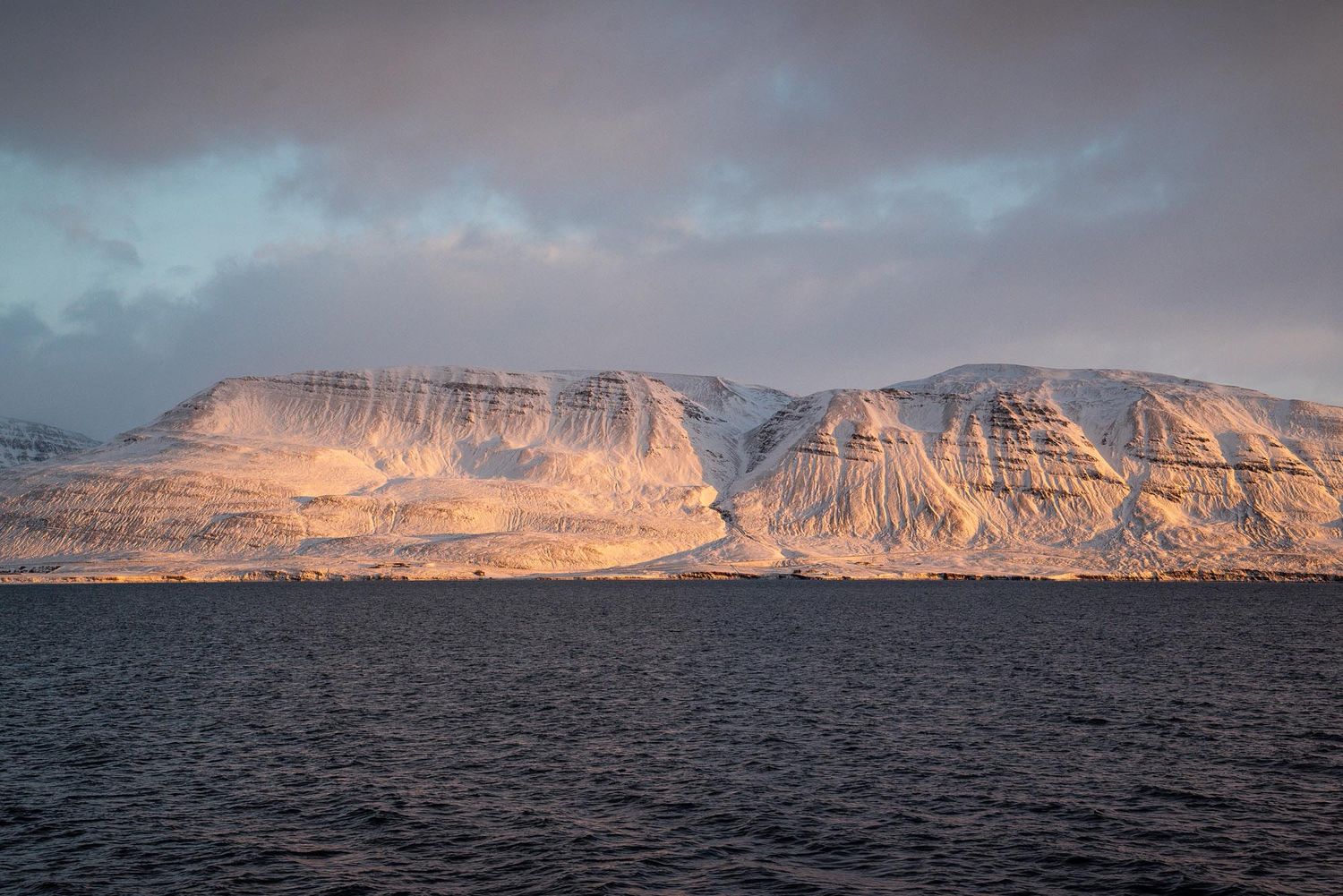 Panoramic view of rugged mountain cliffs reflecting golden sunlight over dark waters at dusk.