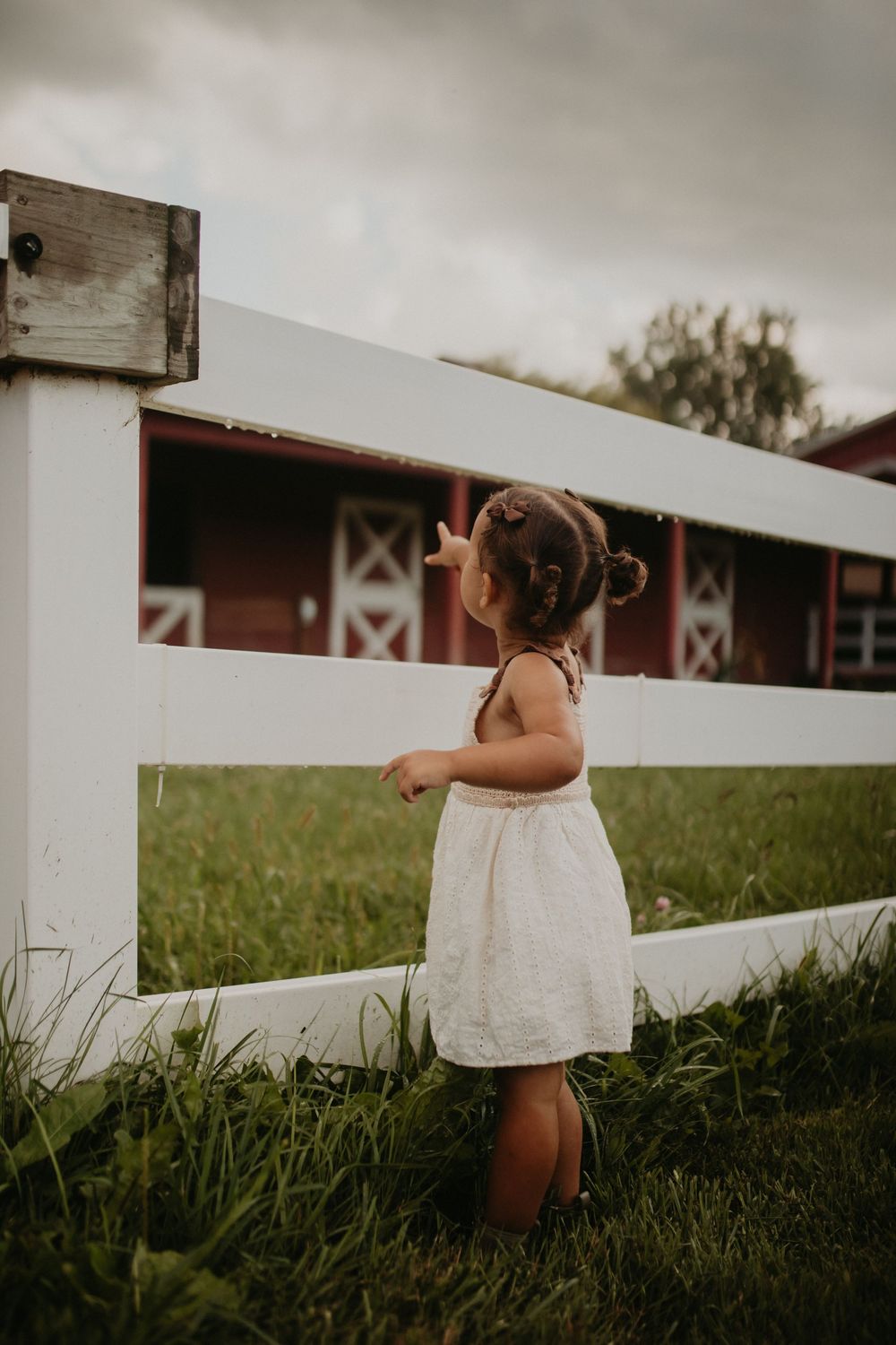 A small child in a white dress walks along a white wooden fence on a grassy hillside in moody lighting.