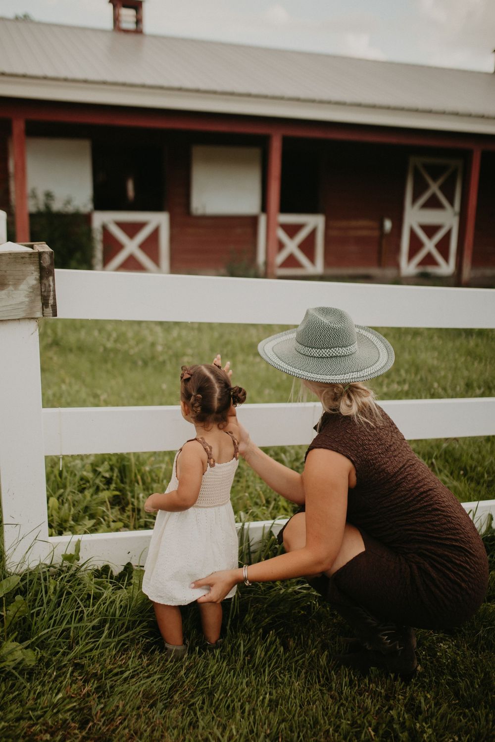 Two people in summer hats lay in green grass near a white fence and red barn, looking at papers together.