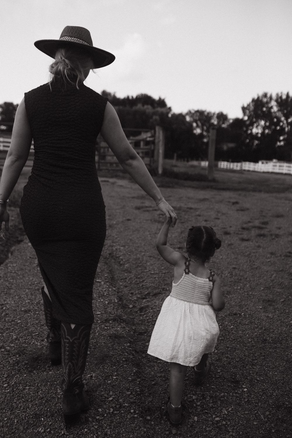 A woman in a black dress and wide-brimmed hat walks hand in hand with a small child in a white dress on a farm.
