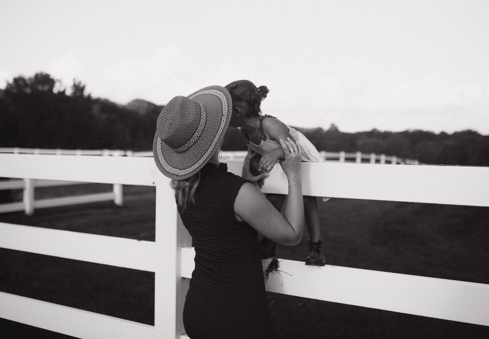 A person leans against a white wooden fence on a farm while wearing a wide-brimmed hat on a sunny day.